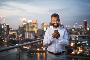 A portrait of businessman with smartphone standing against night London rooftop view panorama. Copy space.