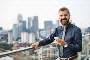 Un retrato de un hombre de negocios con un teléfono inteligente de pie frente al panorama de la vista de la azotea de Londres. Espacio de copia.