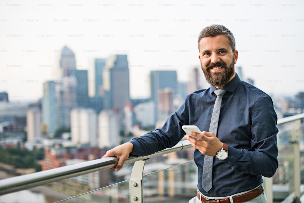 A portrait of businessman with smartphone standing against London rooftop view panorama. Copy space.