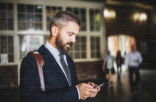 Un hombre de negocios hipster con un teléfono inteligente parado en el interior de la ciudad, enviando mensajes de texto. Espacio de copia.