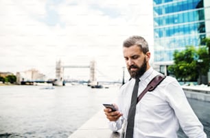 Hombre de negocios hipster serio con teléfono inteligente parado junto al río Támesis en Londres, mensajes de texto. Espacio de copia.