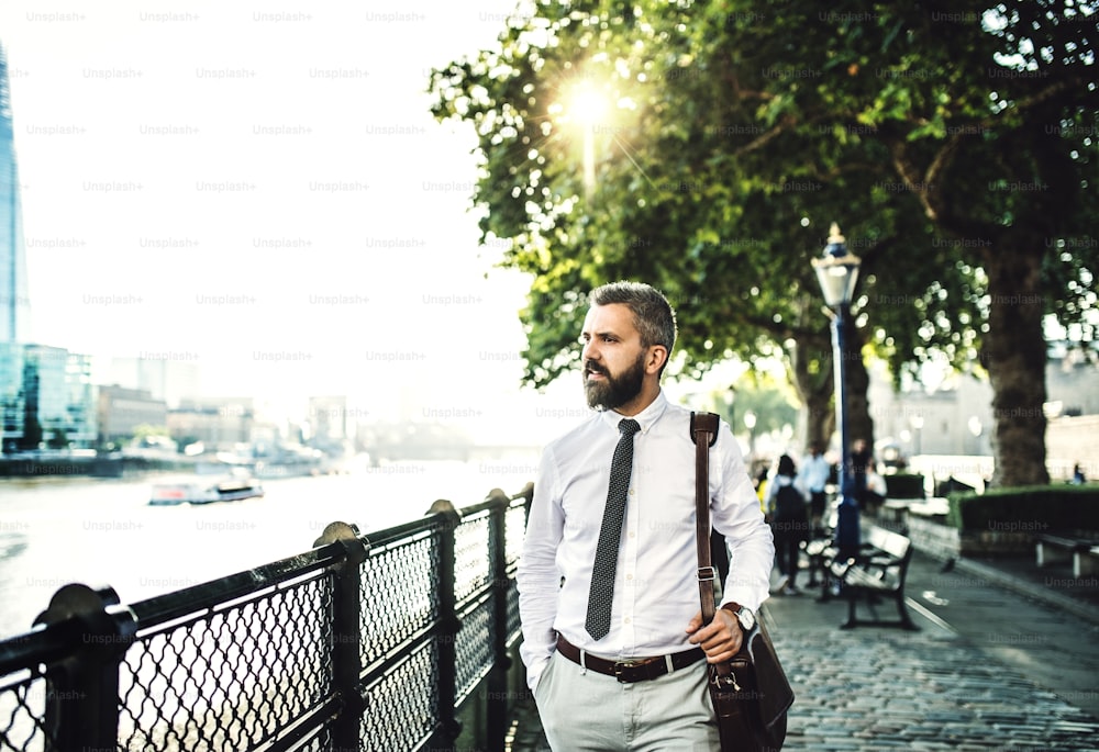 Ernsthafter Hipster-Geschäftsmann mit Laptoptasche, der am Fluss in London spazieren geht.