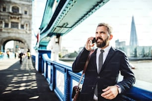 Hipster-Geschäftsmann mit Smartphone steht auf der Brücke in der Londoner City und telefoniert.