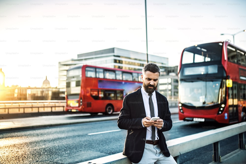 Hipster businessman with smartphone standing by the road in London, text messaaging. Copy space.