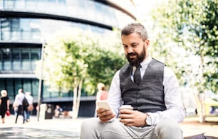 Serious hipster businessman with smartphone and coffee cup sitting outdoors in the city, text messaging.