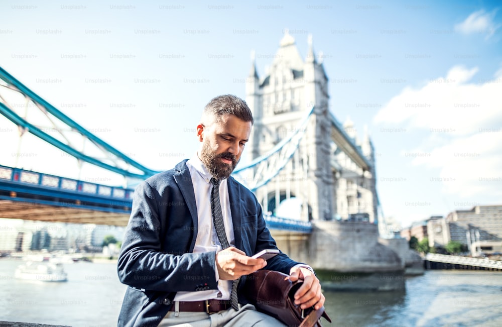 Hipster businessman with a smartphone sitting by the Tower Bridge in London, text messaging. Copy space.
