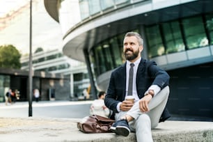 Un portrait d’homme d’affaires hipster avec un sac d’ordinateur portable et une tasse de café assis à l’extérieur dans la ville.