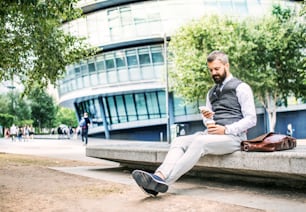 Ernsthafter Hipster-Geschäftsmann mit Smartphone, Laptoptasche und Kaffeetasse, der draußen in der Stadt sitzt, SMS.
