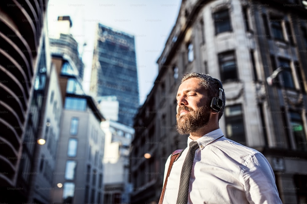 Hipster businessman with headphones standing on the street in London city, listening to music. Copy space.
