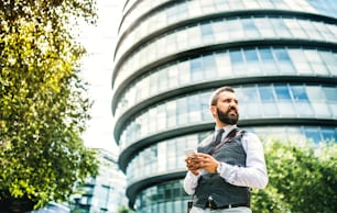 Low angle portrait of businessman with smartphone standing on the street in city in front of a building, text messaging.
