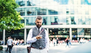 Hipster businessman with smartphone standing on the street in city, text messaging.