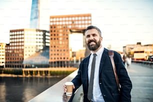 Mature hipster businessman with coffee in a paper cup standing on the bridge in London. Copy space.