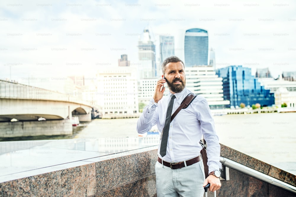 Hipster businessman with smartphone and suitcase standing by the river Thames in London, making a phone call. Copy space.