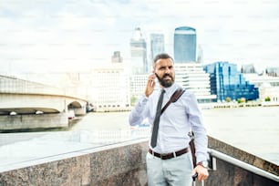 Hipster businessman with smartphone and suitcase standing by the river Thames in London, making a phone call. Copy space.