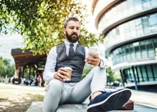 Hipster-Geschäftsmann mit Kaffee und Smartphone sitzt auf einer Bank auf der Straße in London, SMS.