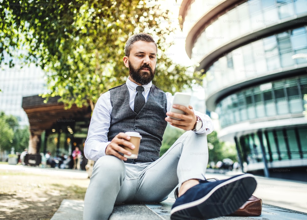 Hipster businessman with coffee and smartphone sitting on a bench on the street in London, text messaging.