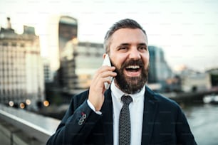 Laughing hipster businessman with smartphone standing on the bridge in the city, making a phone call.