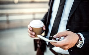 A close-up of an unrecognizable businessman with smartphone and coffee in a paper cup in the city, text messaging. Copy space.