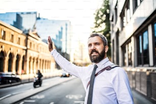 Hipster businessman standing on the street in London, raising his shand to hail a taxi cab.