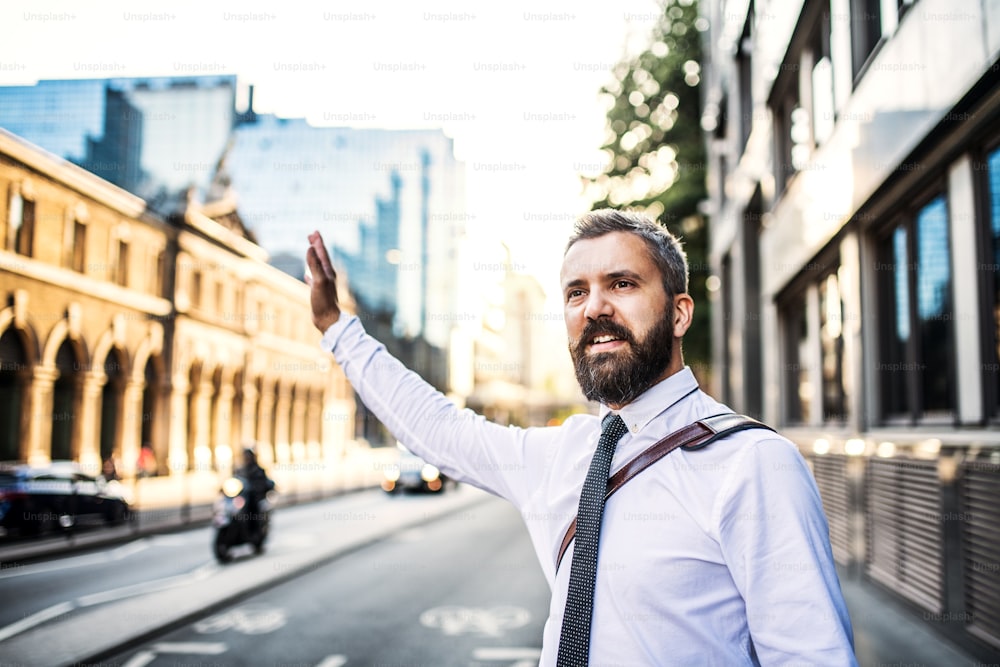 Hipster businessman standing on the street in London, raising his shand to hail a taxi cab.