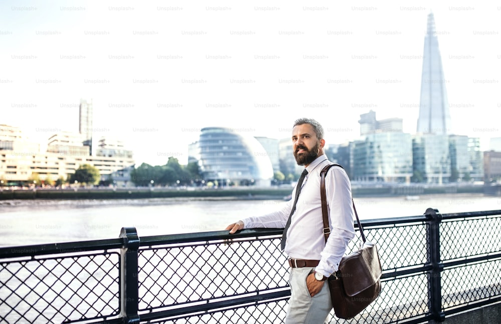 Happy hipster businessman with laptop bag standing by the river in London, holding onto a railing.