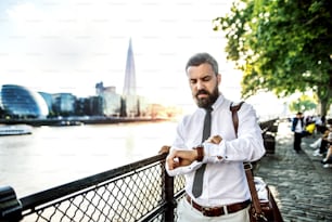 Serious hipster businessman with laptop bag walking by the river in London, checking the time.