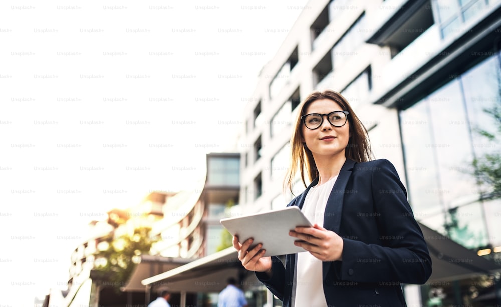 A young businesswoman using tablet outdoors in front of a building in a city. Copy space.