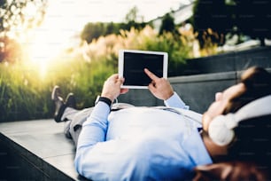 A close-up of a businessman with tablet and headphones, lying down on concrete steps outdoors, a finger on the screen. Copy space.