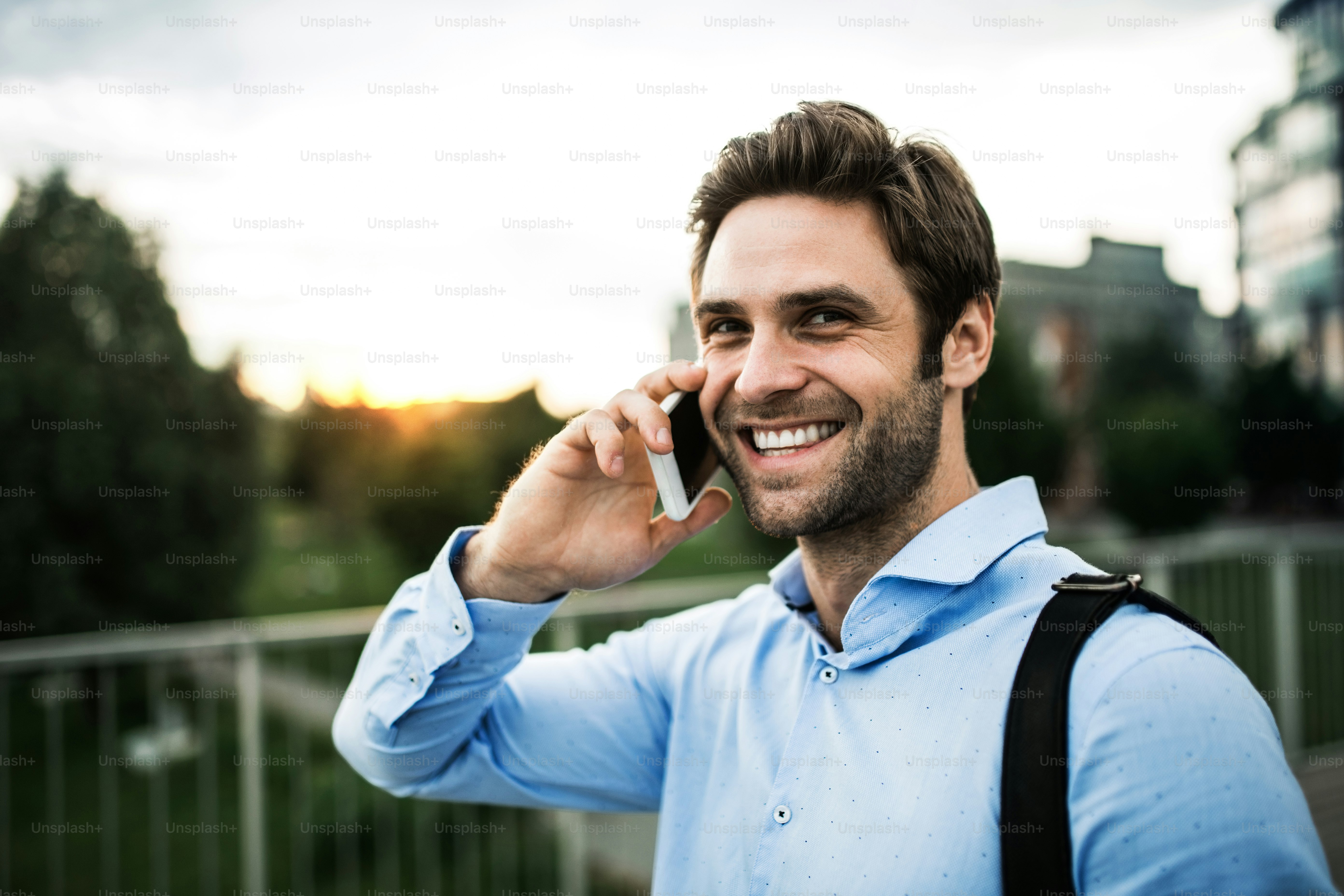 A young businessman with a smartphone walking on a bridge at sunset, making a phone call.