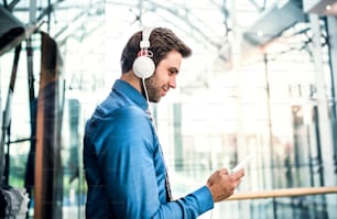 A young businessman with smartphone and headphones standing in a modern shopping center, listening to music.