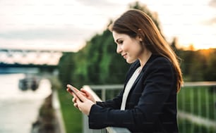 Eine junge Geschäftsfrau steht draußen am Flussufer und benutzt ein Smartphone.
