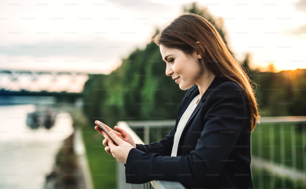 Eine junge Geschäftsfrau steht draußen am Flussufer und benutzt ein Smartphone.