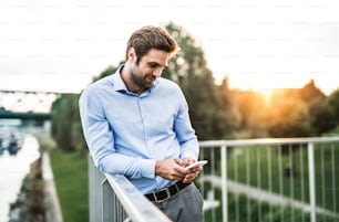 A young businessman with smartphone standing on a bridge at sunset, leaning on a railing. Copy space.