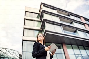 A low angle view of young businesswoman using tablet outdoors in front of a building in a city. Copy space.