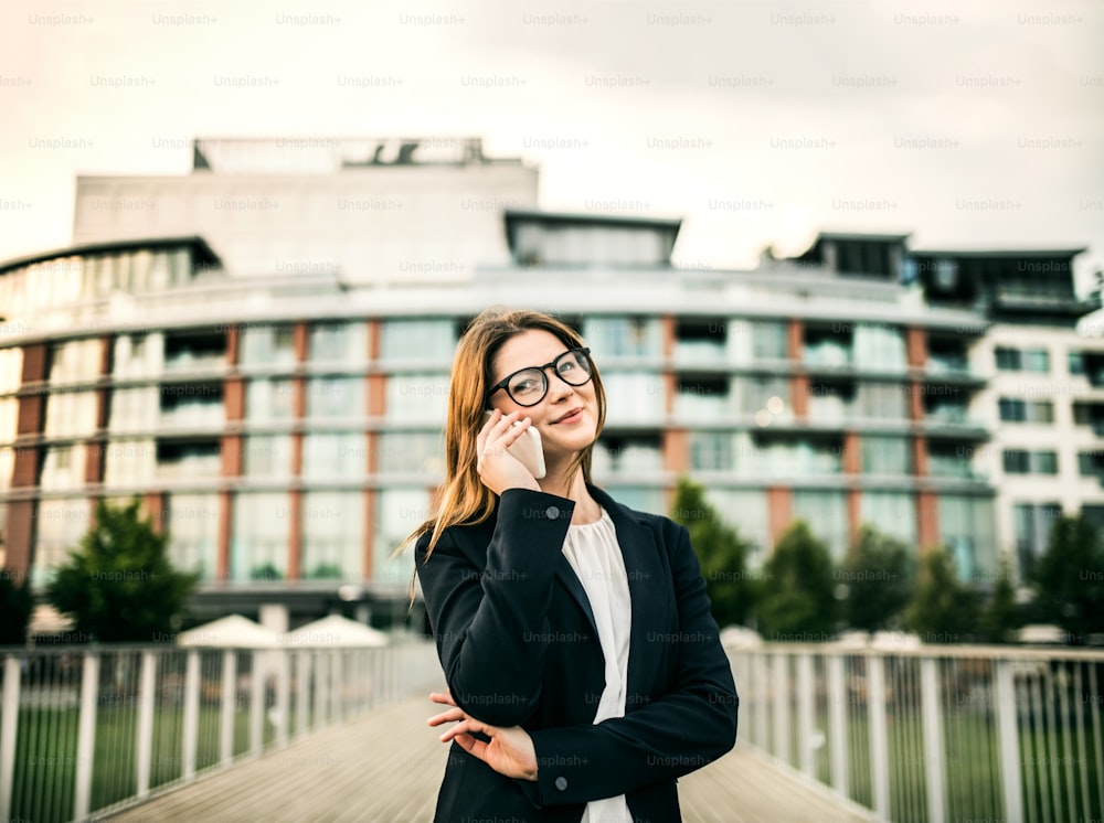 A happy young businesswoman with glasses and smartphone standing on the bridge in a city, making a phone call.