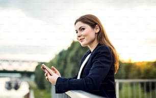 A young businesswoman standing outdoors on the river bank, using smartphone.