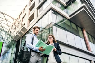 A young businessman and businesswoman standing in front of a building, looking at notes and talking.