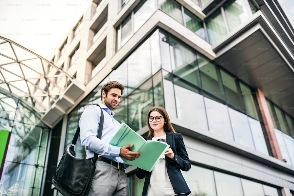Un giovane uomo d'affari e una donna d'affari in piedi di fronte a un edificio, guardando appunti e parlando.