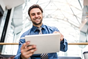 A young businessman with tablet and headphones standing in a modern building, listening to music.