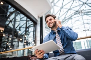 A young businessman with tablet and headphones standing in a modern building, listening to music.