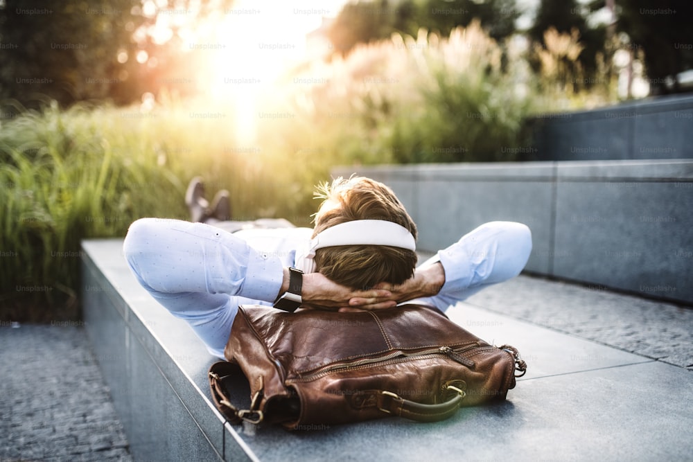 A businessman with headphones, lying on the steps outdoors at sunset, a head resting on a laptop bag.