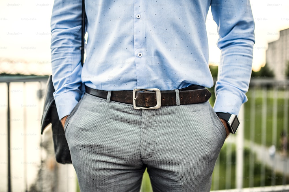 An unrecognizable young businessman with a bag and a leather belt walking on a bridge, hands in pockets.