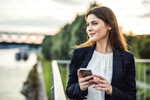 A young businesswoman standing outdoors on the river bank, using smartphone.