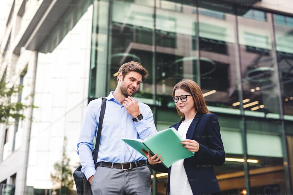 A young businessman and businesswoman standing in front of a building, looking at notes and talking.