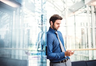 Un jeune homme d’affaires avec smartphone debout dans un bâtiment moderne, messagerie texte. Espace de copie.