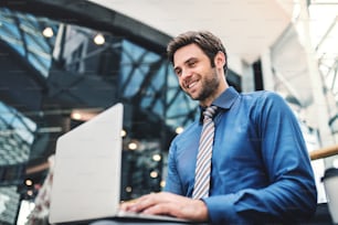 A young handsome businessman sitting on a bench in a modern building, using laptop.