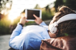 A close-up of a businessman with tablet and headphones, lying down on the bench outdoors, a finger on the screen. Copy space.