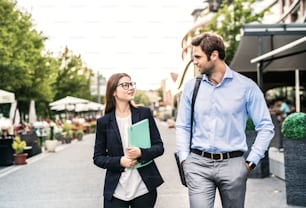 A young businessman and businesswoman walking on a sidewalk, talking.