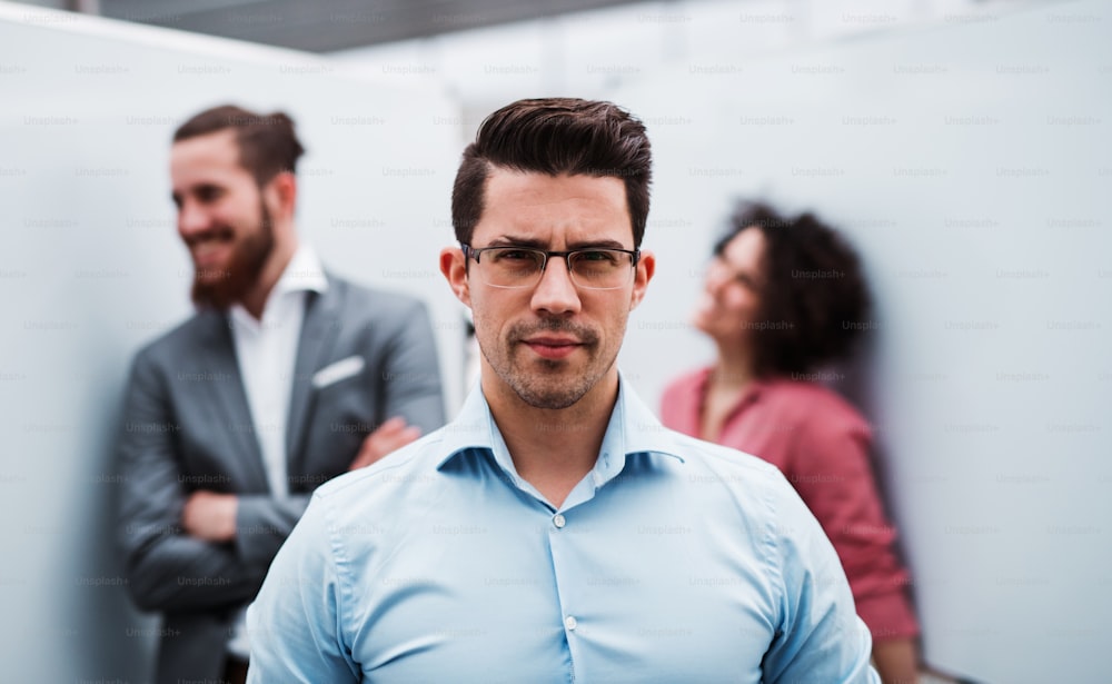 A portrait of young businessmanstanding in office, colleagues in the background.