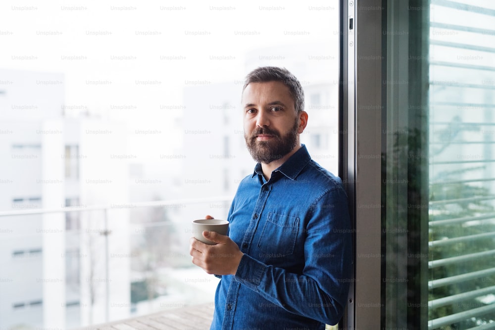 A man standing by the window, holding a cup of coffee at home or in an office.